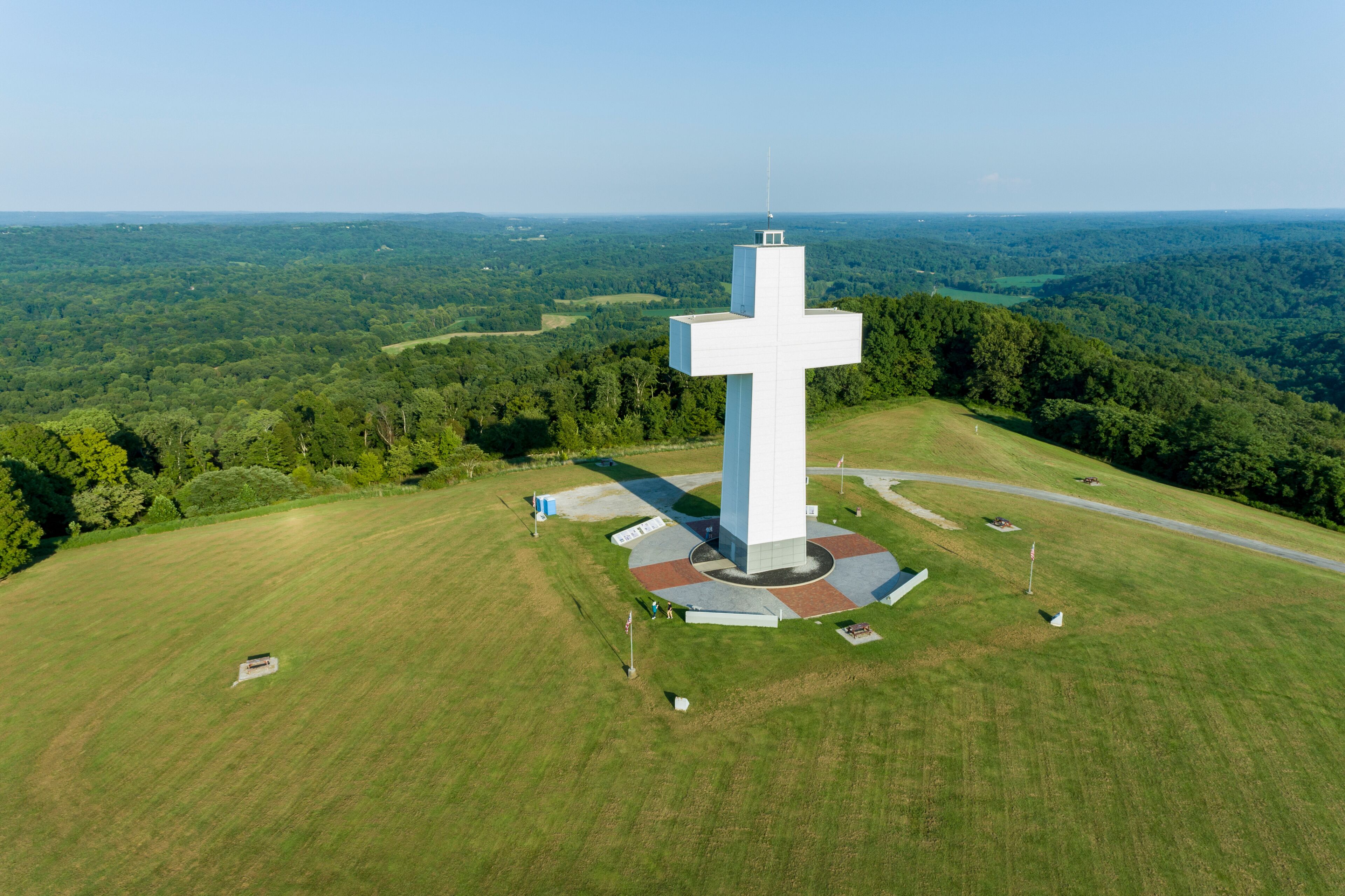 Un grand monument en forme de croix blanche, entouré d’une place circulaire, se dressant sur un paysage verdoyant et luxuriant, avec des collines qui se dessinent à l’horizon.