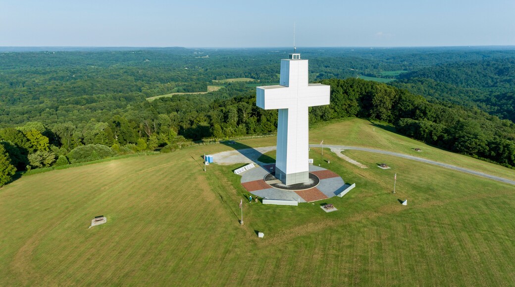 Un grand monument en forme de croix blanche, entourĂ© dâune place circulaire, se dressant sur un paysage verdoyant et luxuriant, avec des collines qui se dessinent Ă lâhorizon.