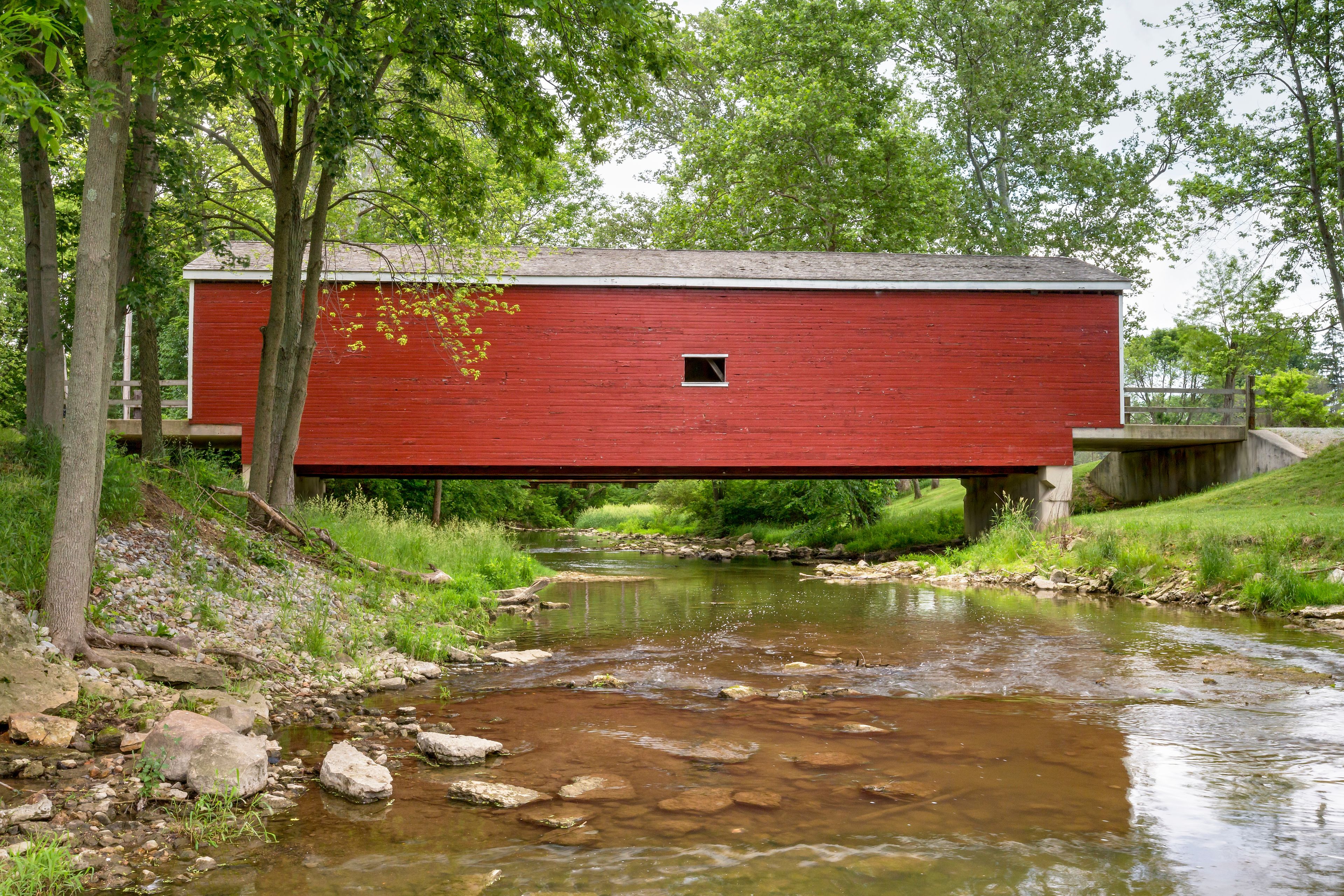 A red wooden structure, possibly a bridge, over a shallow stream with rocks and greenery around.
