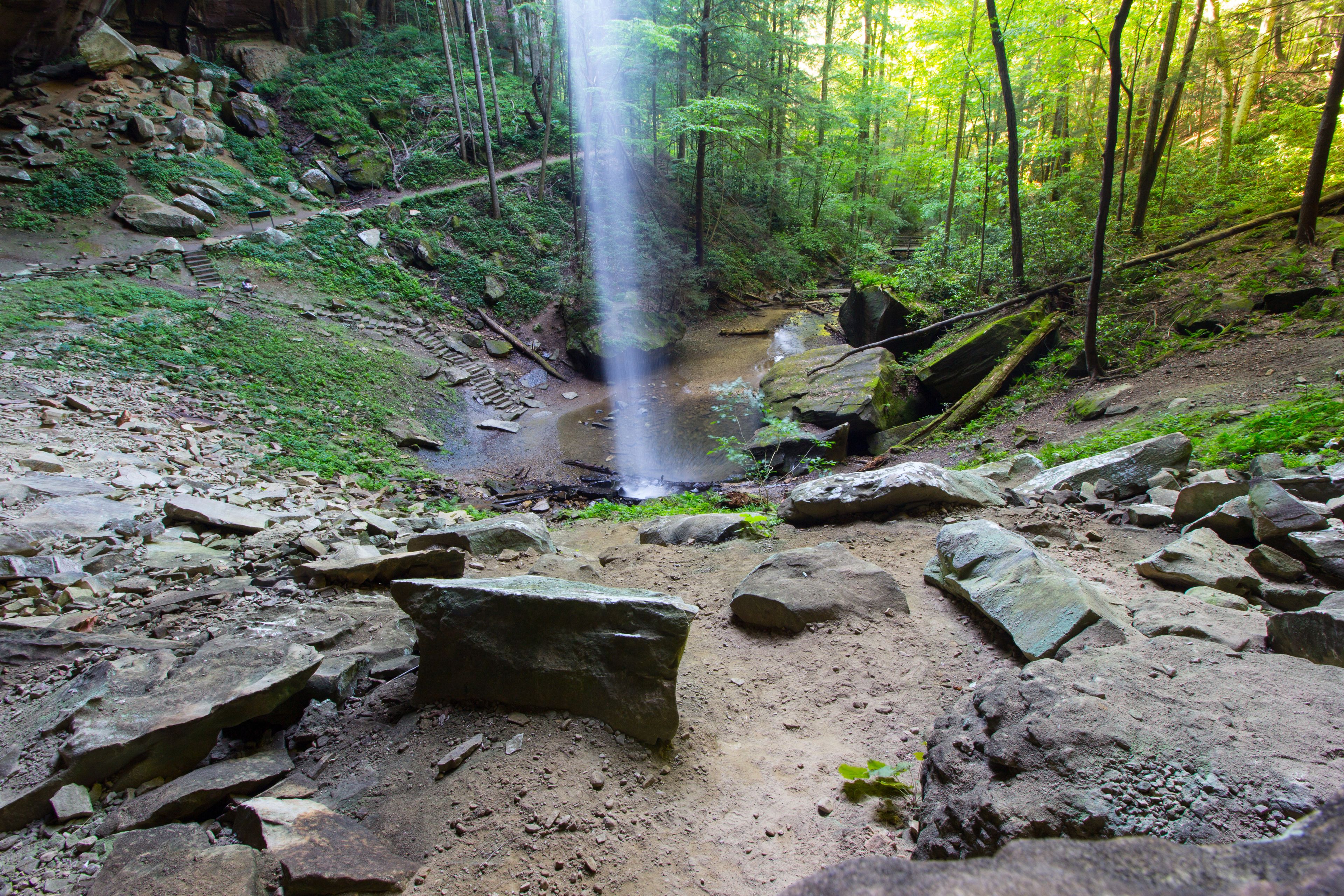 Une cascade située dans une zone boisée, avec des rochers et un sentier de terre.