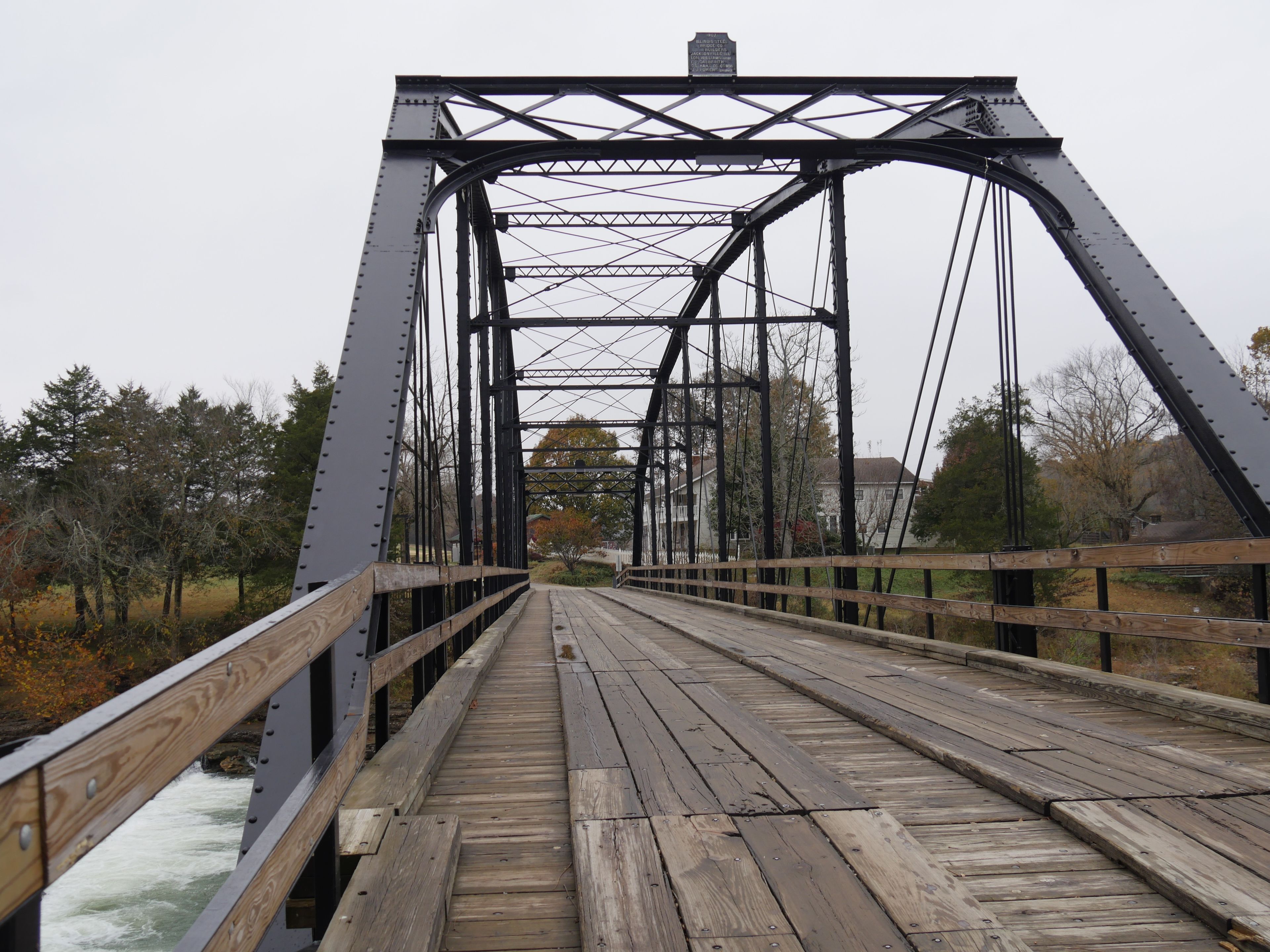 A wooden bridge with metal trusses over a river, surrounded by trees and houses.