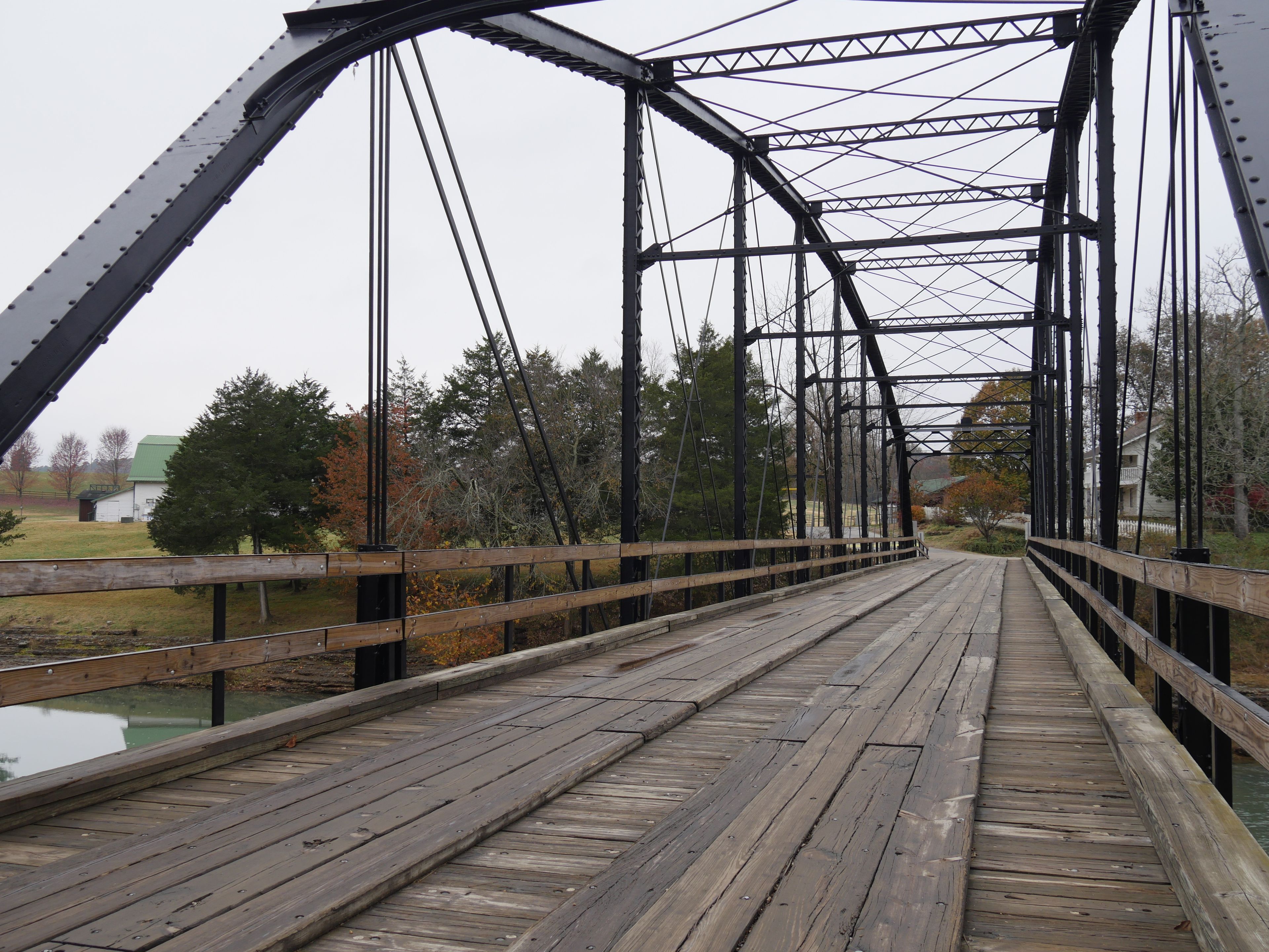 A wooden bridge with metal trusses, surrounded by trees and a grassy area.