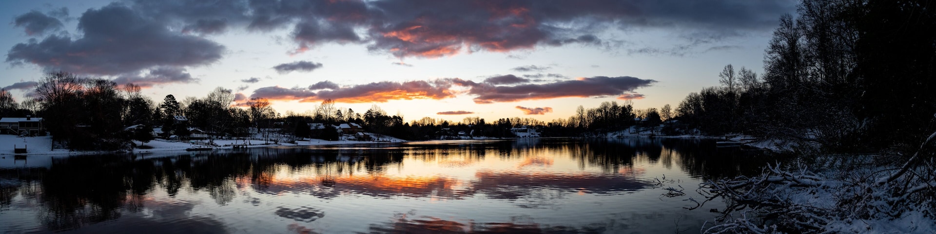 A serene lake at sunset with snow-covered trees and houses in the background.
