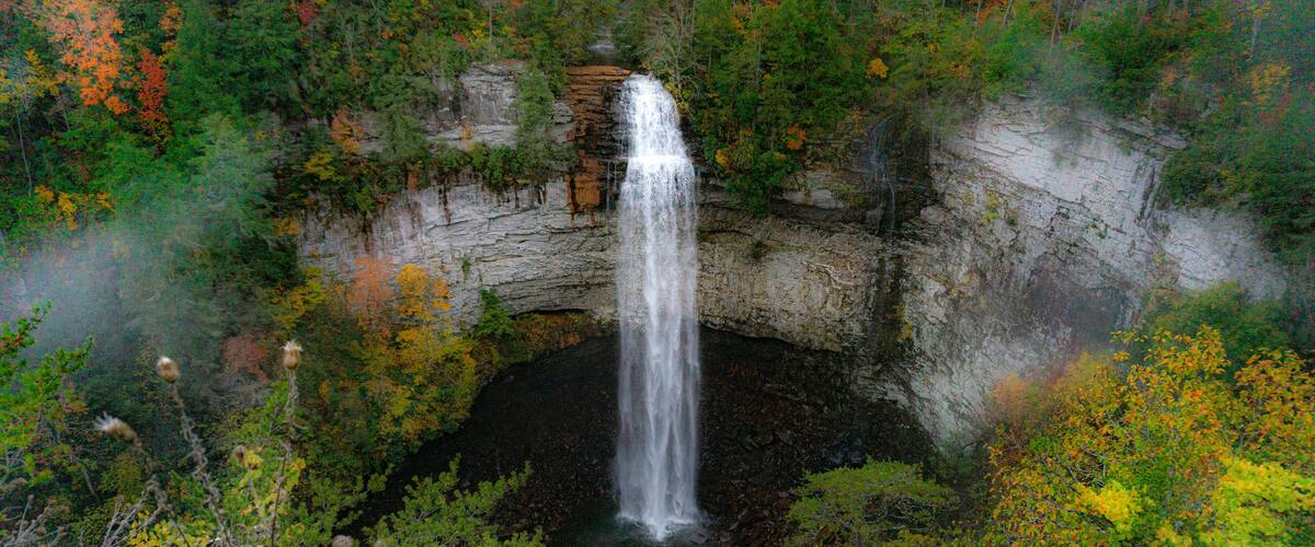 Falls Creek Falls is surrounded by vibrant fall colors and a misty morning haze. The waterfall cascades from a rocky cliff into a dark pool below. Photographed at Falls Creek Falls State Park in TN