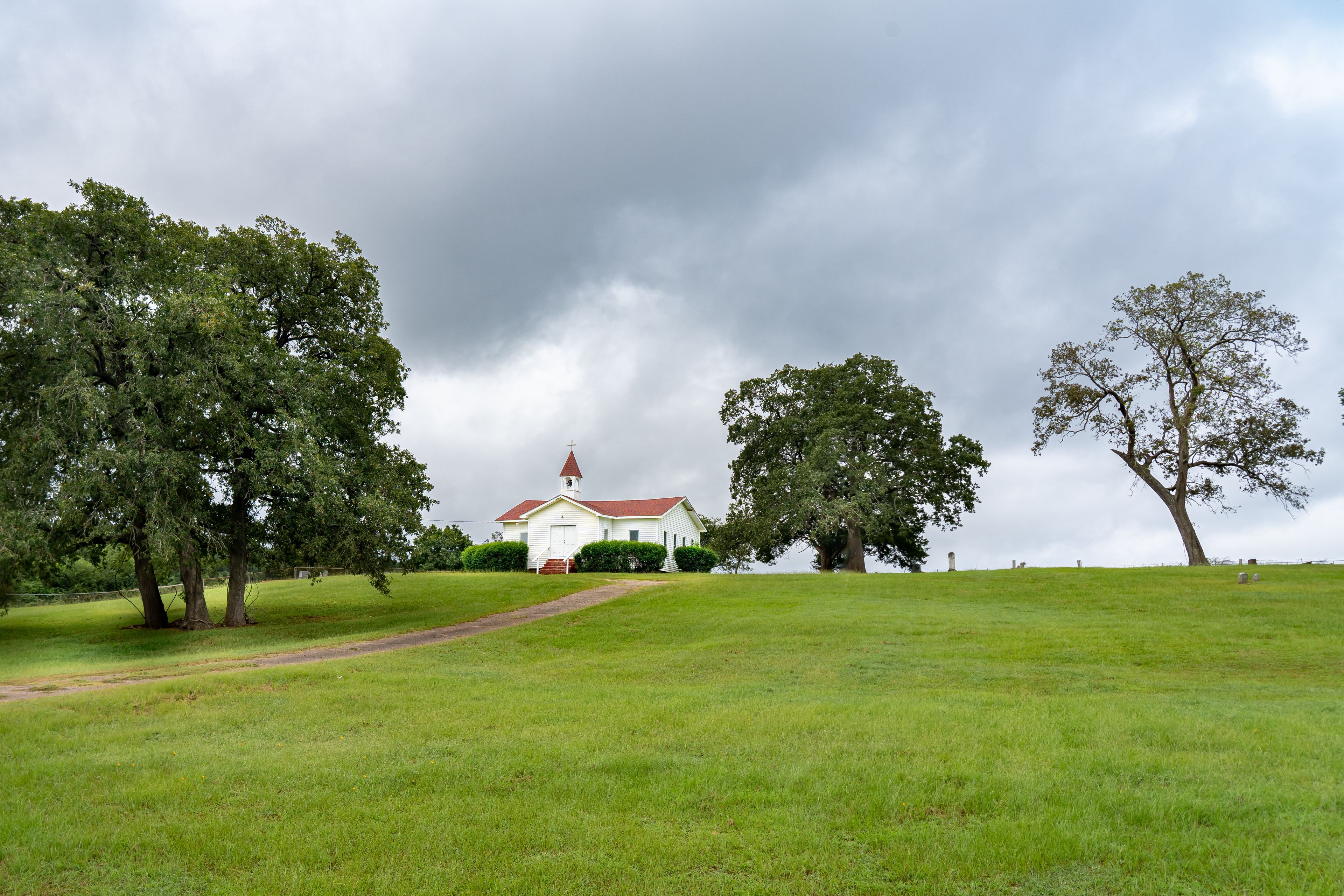 A white church with a red roof on a grassy hill under a cloudy sky.