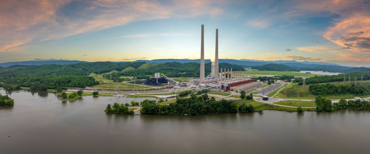 A power plant with multiple smokestacks, surrounded by a lake and forested hills.