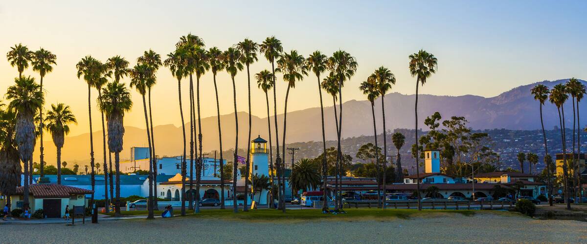A coastal cityscape with palm trees, a lighthouse, and mountains in the background.