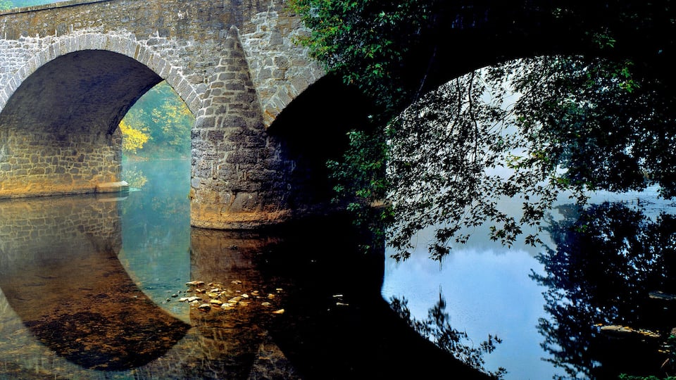 A stone bridge with multiple arches over a calm river, surrounded by trees and greenery.