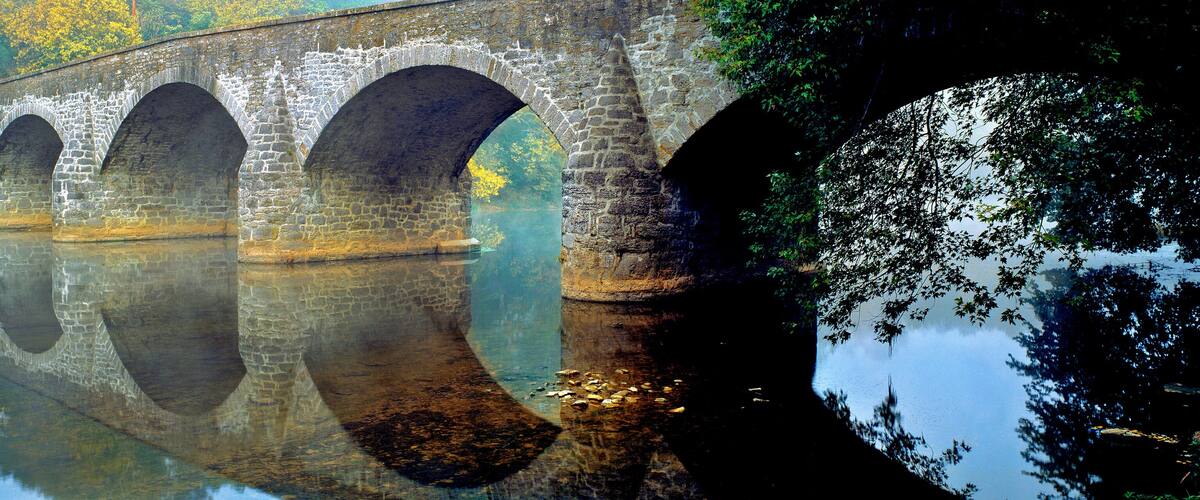 A stone bridge with multiple arches over a calm river, surrounded by trees and greenery.