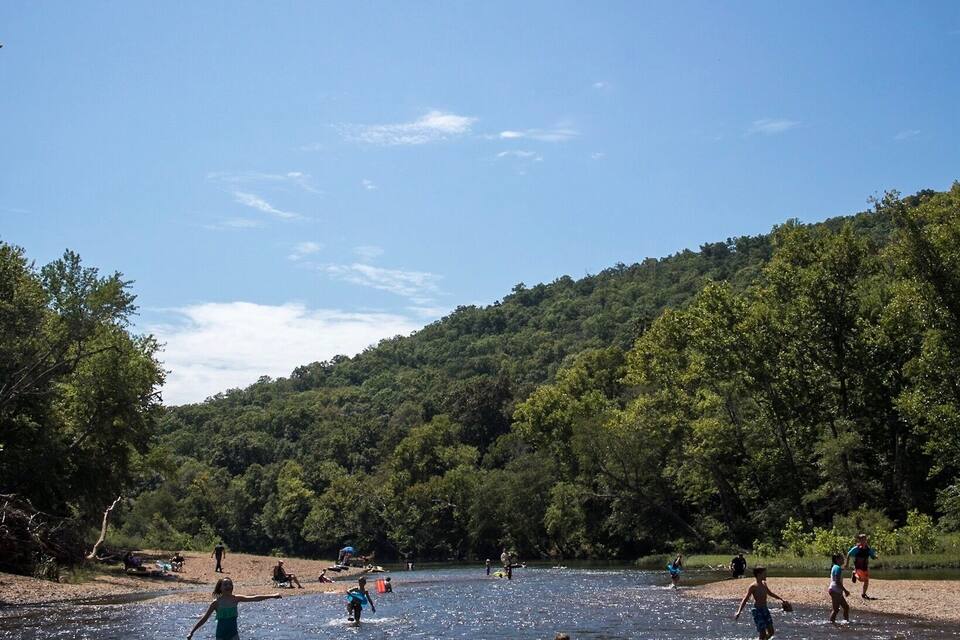 A river with people swimming and standing in the water, surrounded by a forested hillside.