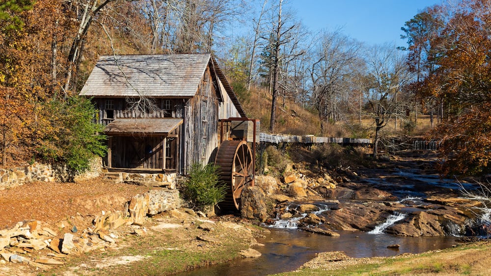 Un moulin en bois, doté d’une roue à eau, se trouve au bord d’une rivière, entouré d’arbres aux couleurs automnales.