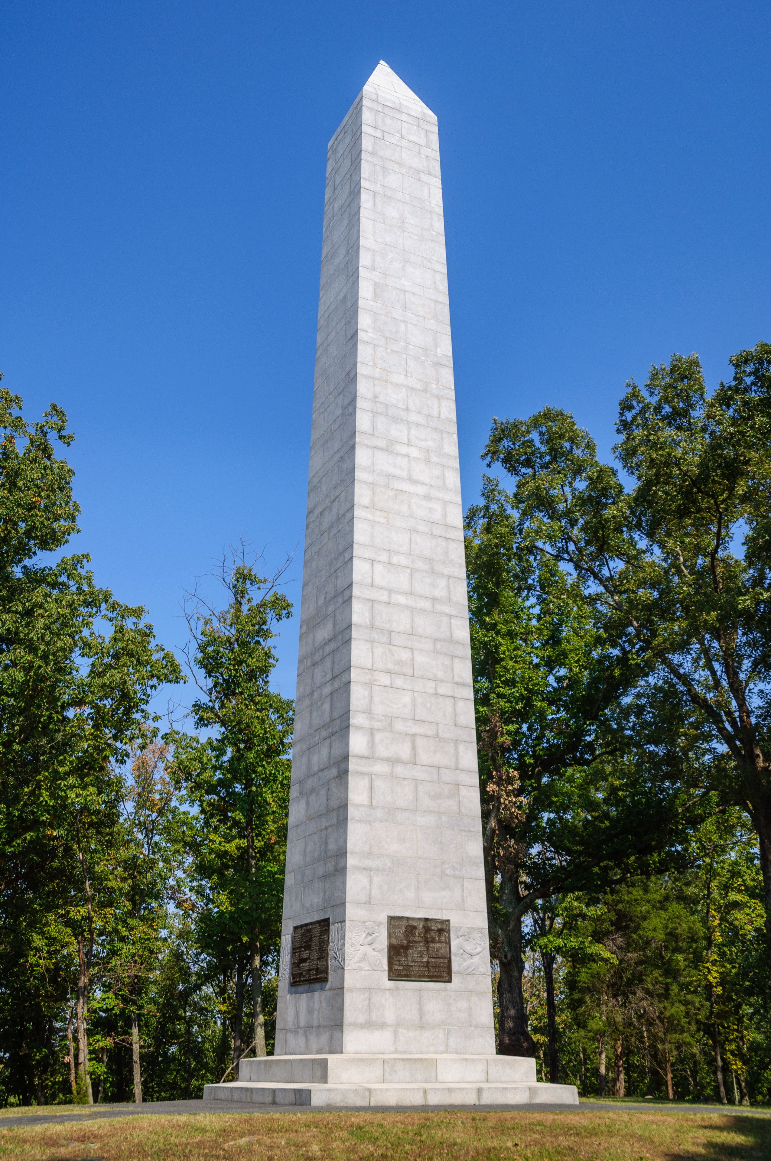 A tall, white obelisk monument with inscriptions, surrounded by trees and grass.