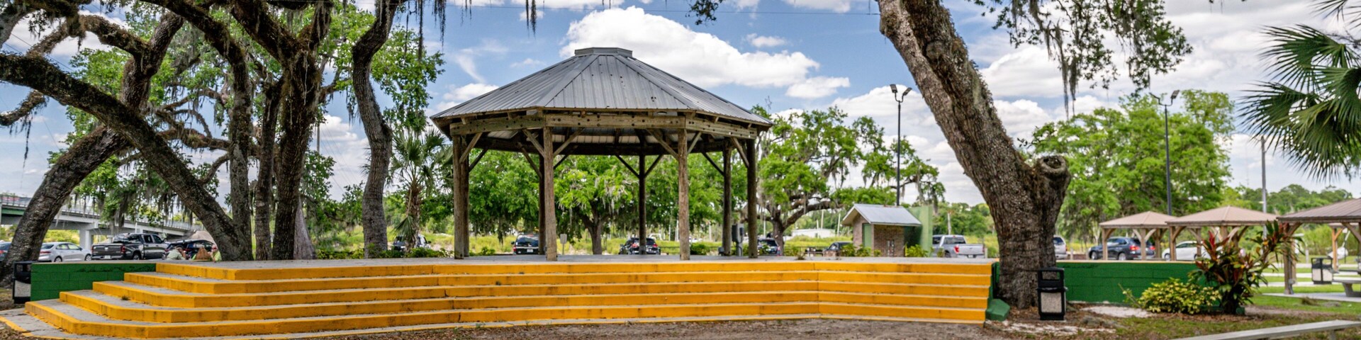 Ein Gazebo mit Dach, umgeben von gelben Stufen und Bänken, unter großen Bäumen mit hängenden Moosen.
