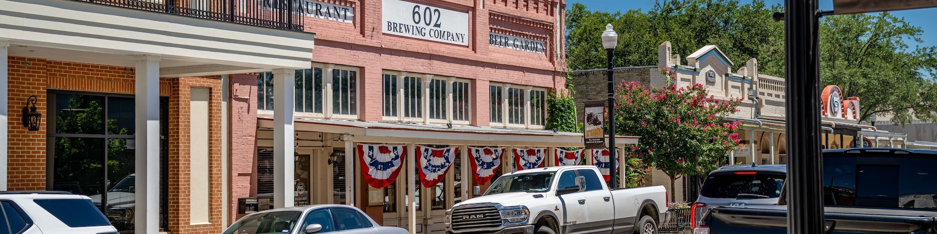 Una calle con un edificio de ladrillo identificado como '602 BREWING COMPANY', una camioneta pickup blanca y un automóvil plateado.