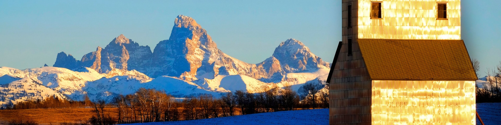 A wooden structure with a sloped roof, a snow-covered field, and a mountain range in the background.