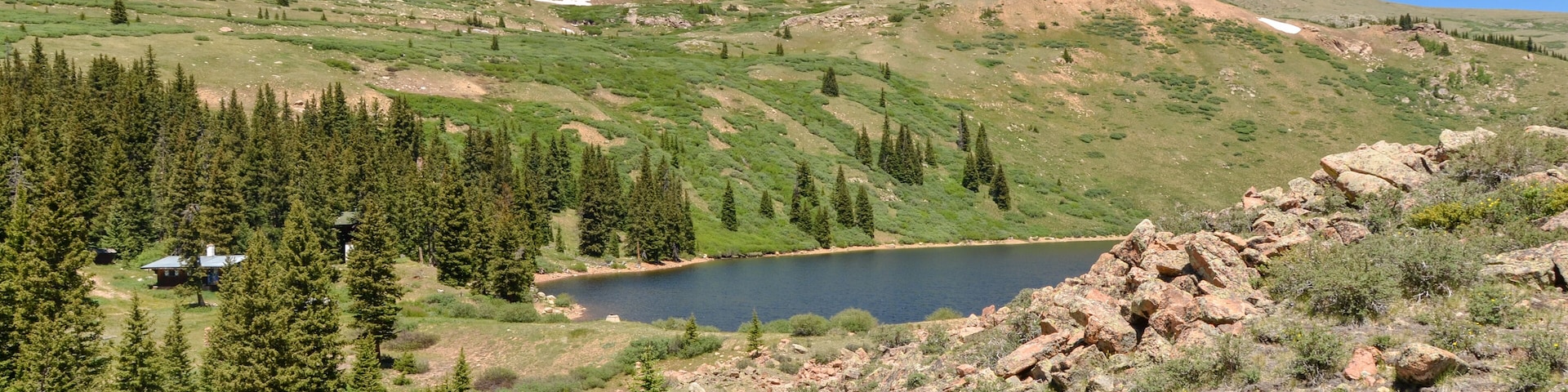 A mountainous landscape with a lake, forested areas, and a small cabin.