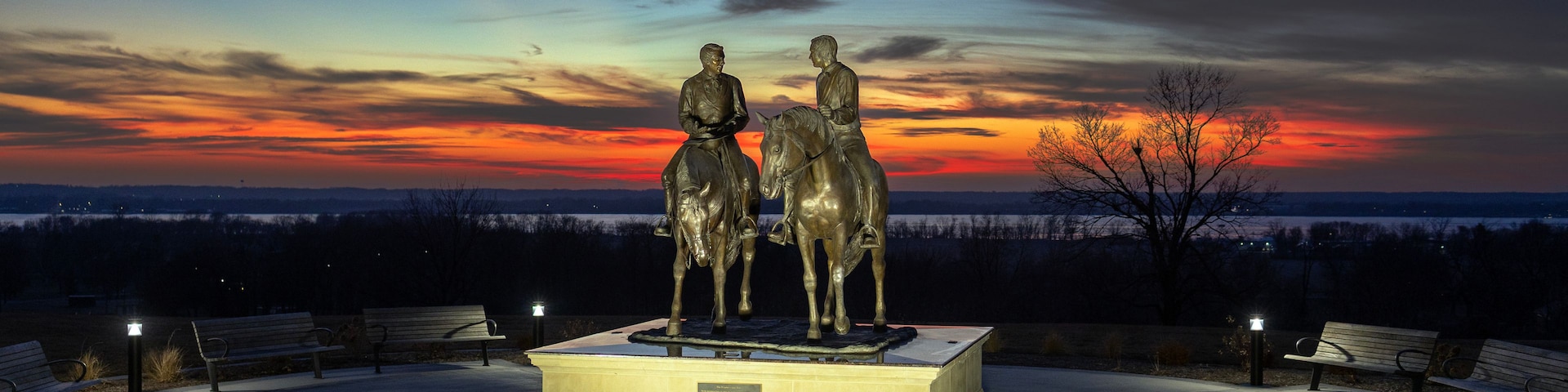 Una estatua de dos figuras a caballo, con un atardecer de fondo y bancos alrededor.