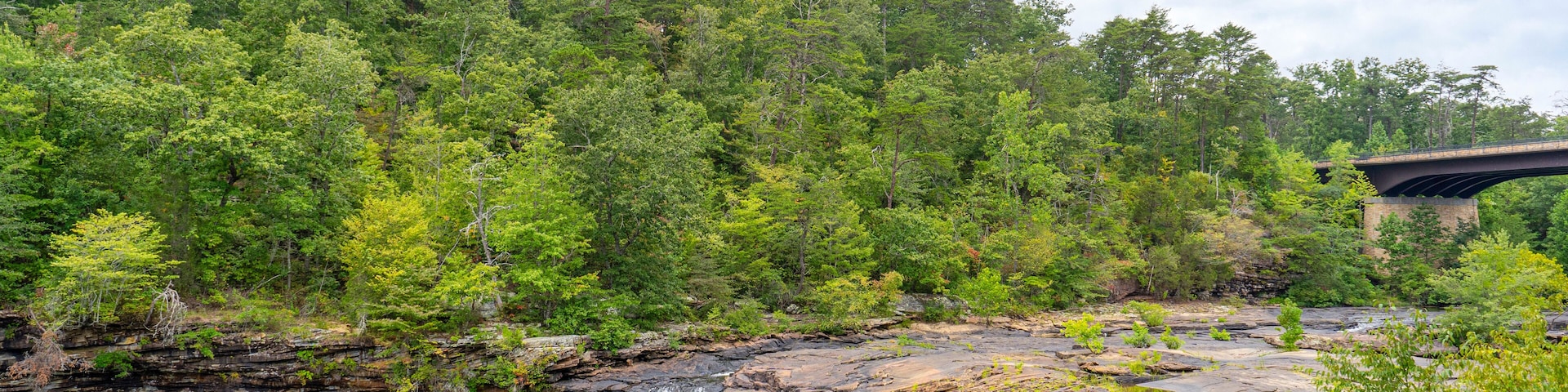 In the summer, Little River Falls in Little River Canyon National Preserve in Alabama is surrounded by green forests set against a cloudy blue sky. The Hwy 35 bridge is visible in the background.