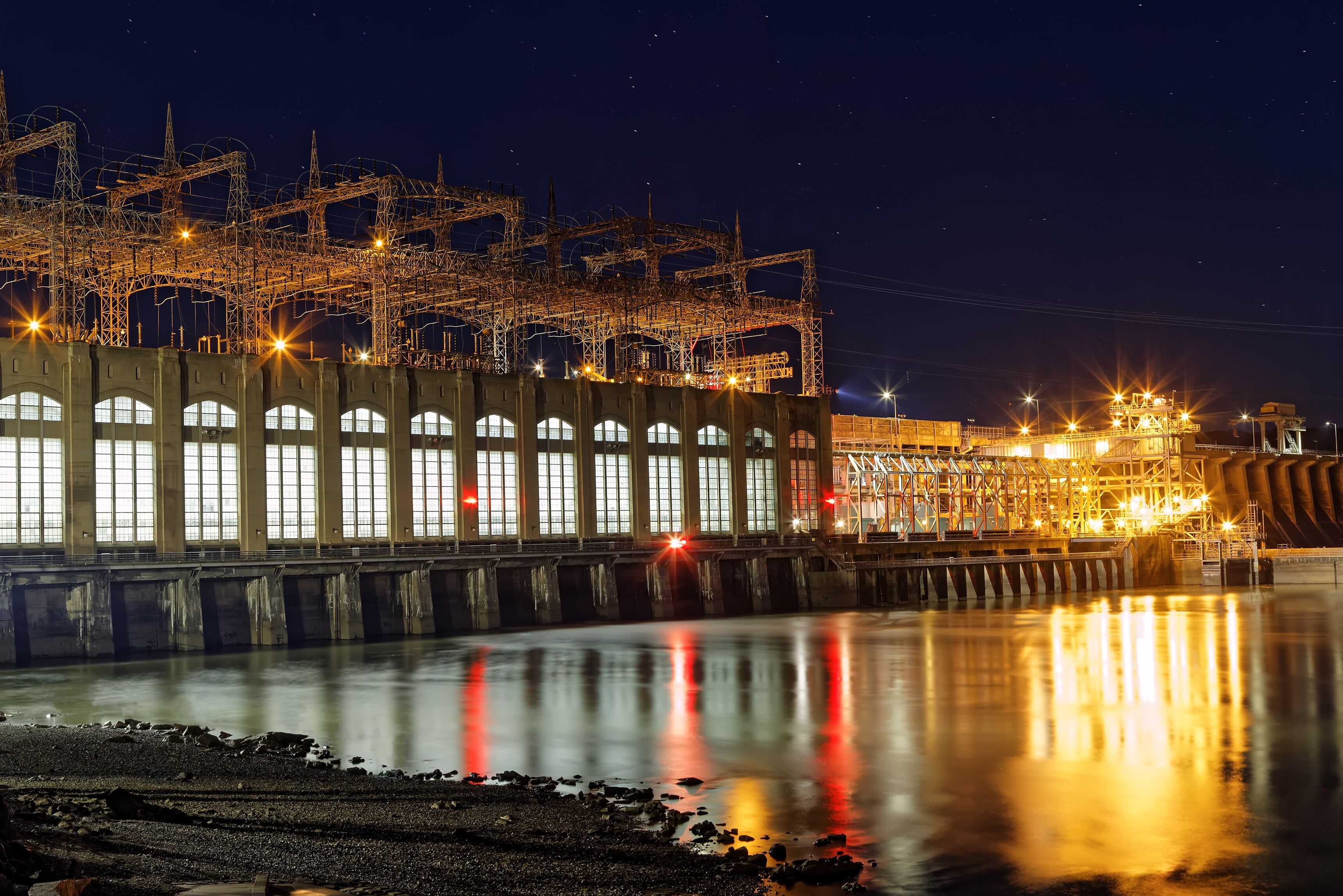 A nighttime industrial complex with illuminated structures and a calm river reflecting lights.