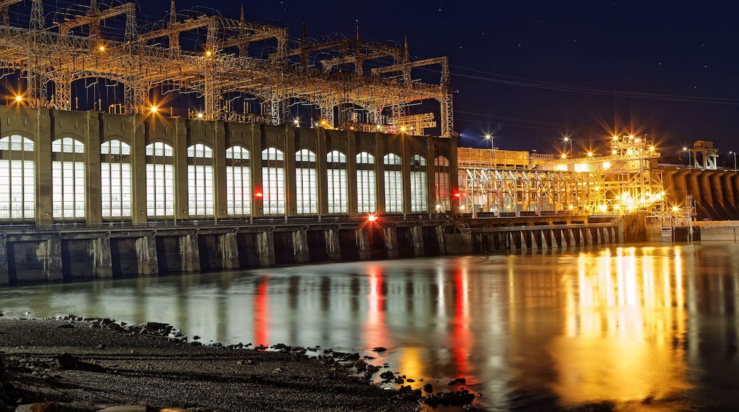 A nighttime industrial complex with illuminated structures and a calm river reflecting lights.