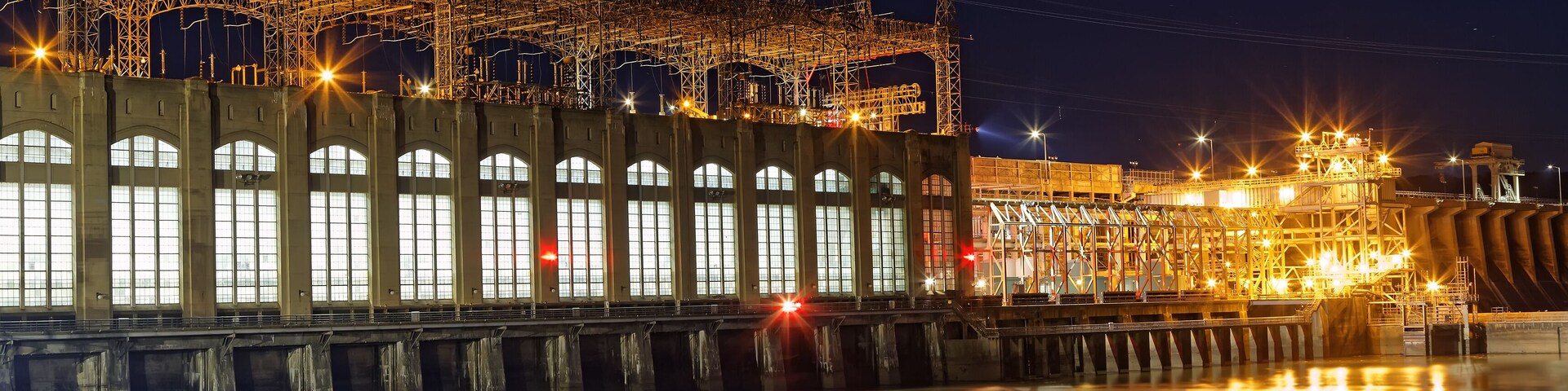 A nighttime industrial complex with illuminated structures and a calm river reflecting lights.