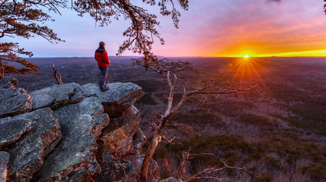 A person standing on a rocky outcrop overlooking a vast landscape during sunset.