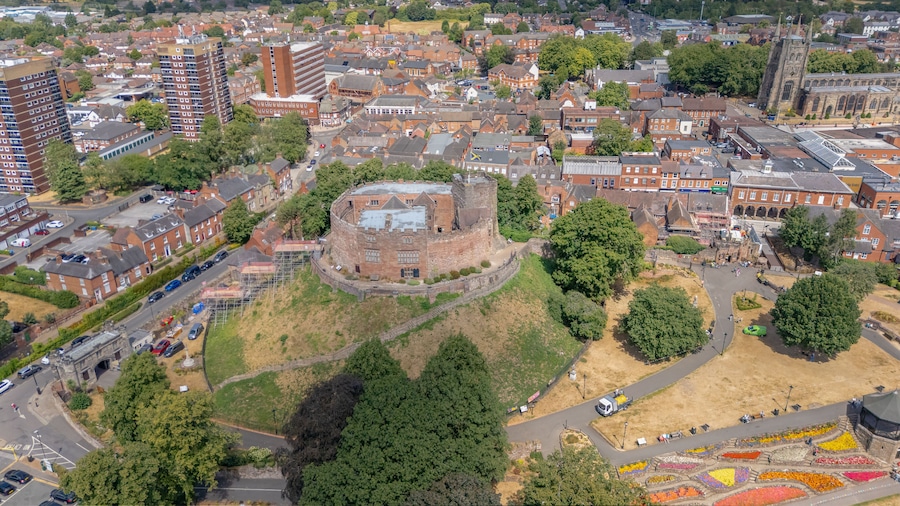 Eine Vogelperspektive zeigt eine historische Burg, umgeben von modernen Gebäuden, einem Parkplatz und einem Blumengarten.