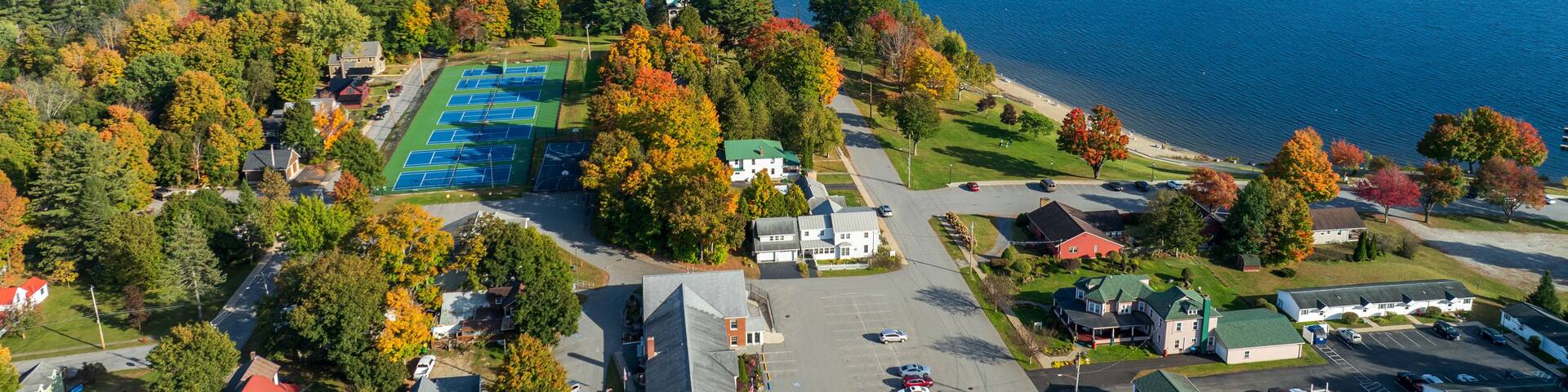 Aerial view of Schroon Lake, New York, showcasing the calm lake, fall foliage, and small town structures nestled along the shoreline under a clear sky.
