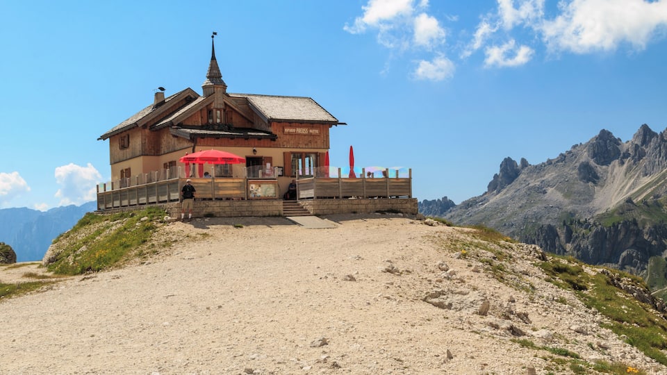 A wooden mountain hut with a red umbrella, surrounded by rocky terrain and mountains.