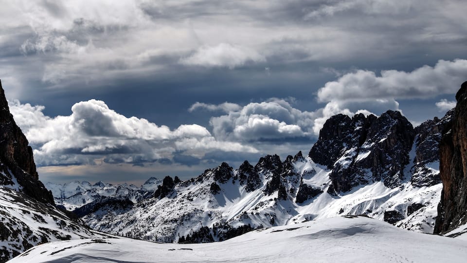 A snow-covered mountain range with rocky peaks and a cloudy sky.