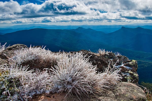 A mountainous landscape with snow-covered vegetation and a distant range of mountains under a partly cloudy sky.