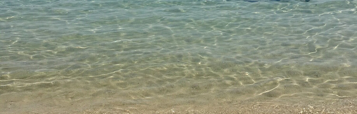 Uma praia com águas cristalinas, pegadas na areia e barcos ao longe.