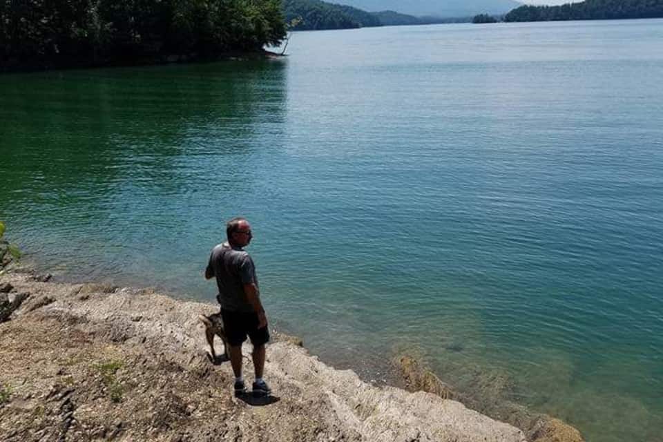 A man standing on a rocky shore overlooking a calm lake with a boat in the distance and mountains in the background.