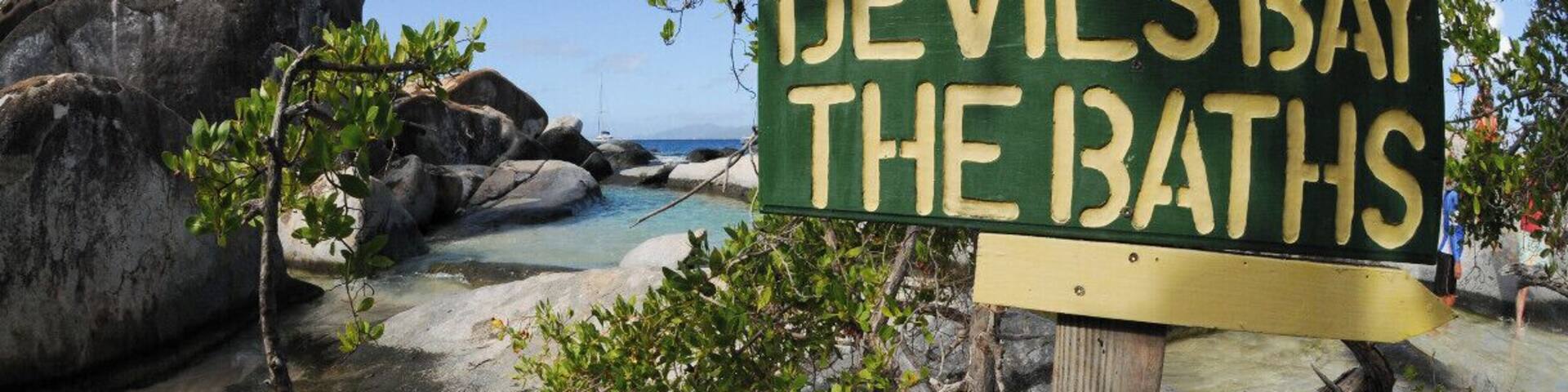 A sign reading "Devil's Bay The Baths" with a beach and rocky landscape in the background.