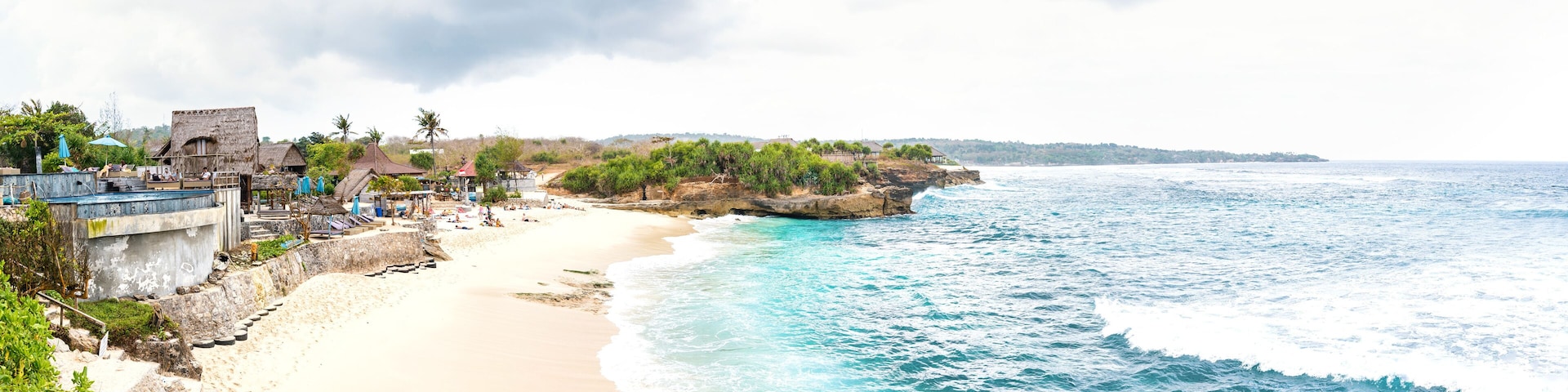 Uma praia com águas turquesa cristalinas, uma faixa de areia e um afloramento rochoso com edifícios.