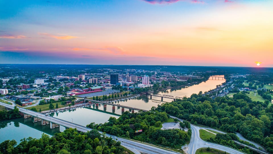 A cityscape at sunset with a river, bridges, and surrounding greenery.