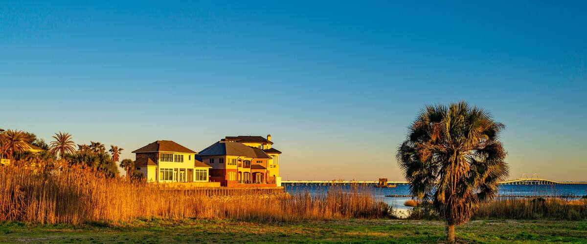 Pensacola Bay Bridge and beach houses along the coastal road, tranquil scenic landscape at golden sunrise with palm tree and dried beach reed plants waving in the wind in Florida, USA