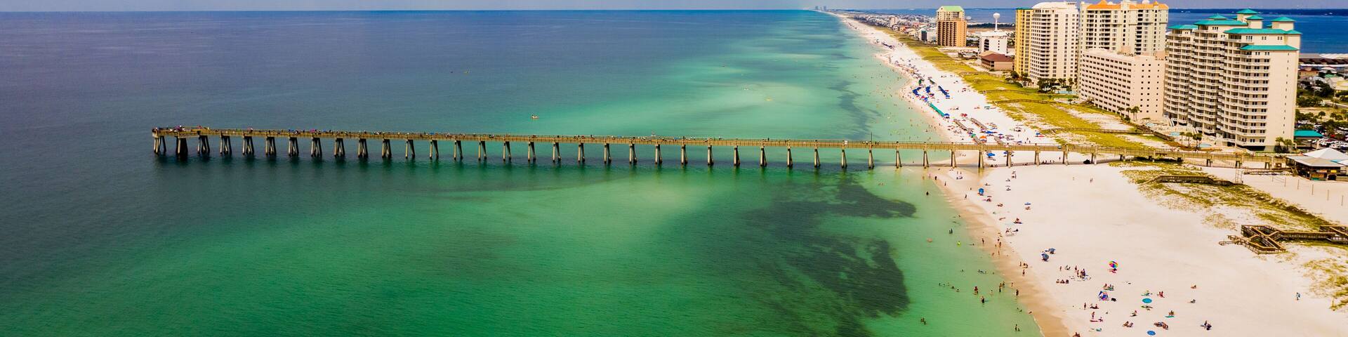 A coastal city with a pier extending into turquoise waters, a sandy beach, and high-rise buildings.