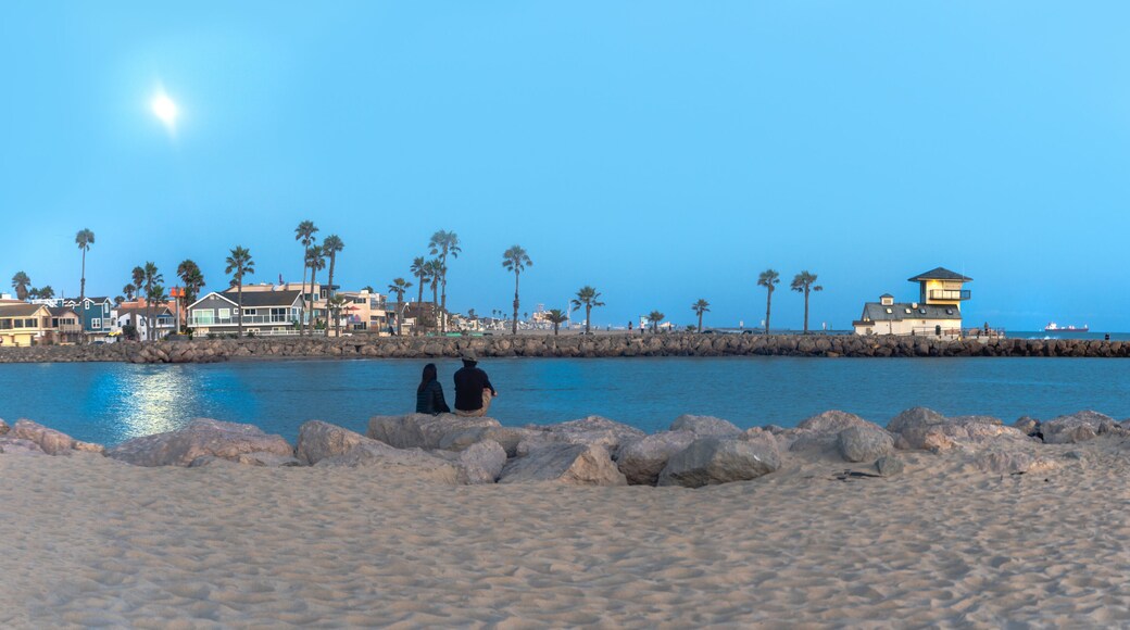 Couple enjoying the moon's reflection on the ocean sitting on the beach looking over at rocky pier with a white building beacon and container ship in background at night time