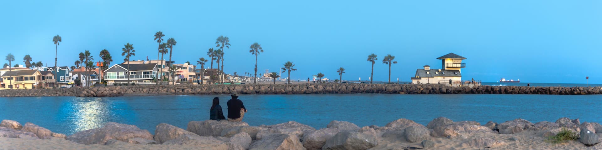 Couple enjoying the moon's reflection on the ocean sitting on the beach looking over at rocky pier with a white building beacon and container ship in background at night time