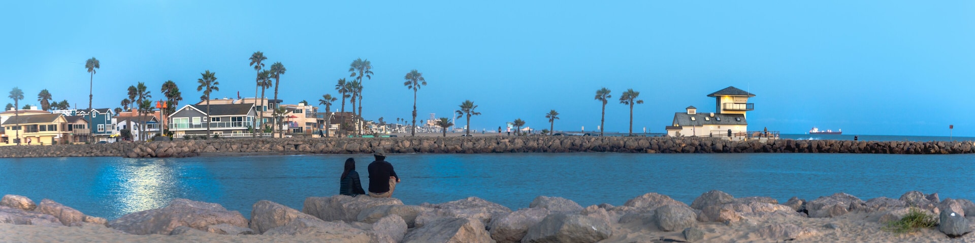 Couple enjoying the moon's reflection on the ocean sitting on the beach looking over at rocky pier with a white building  beacon and container ship in background at night time