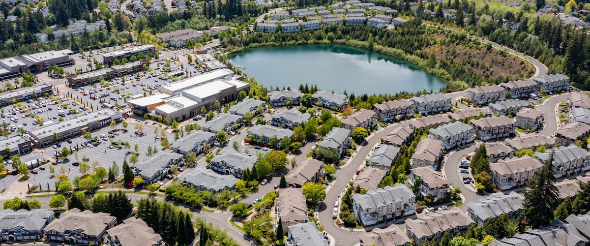 Aerial drone view of Beaverton, Oregon, featuring Progress Lake Park, residential neighborhoods, shopping centers, and parking lots, with tree-lined streets and a mix of suburban and commercial spaces