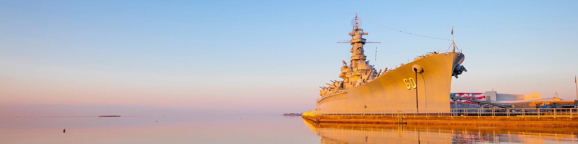 A large naval ship docked at a pier with calm waters reflecting its structure.