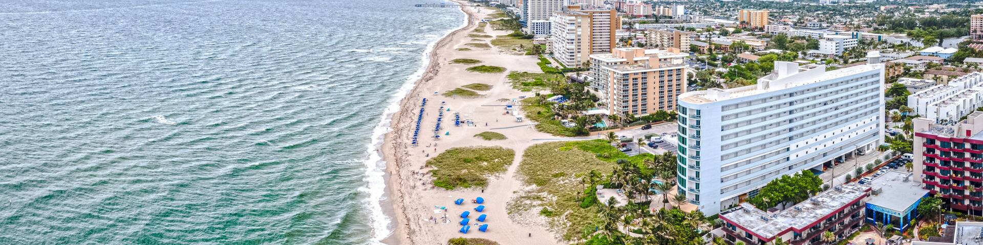 A coastal cityscape with a beach, hotels, and a pier extending into the ocean.