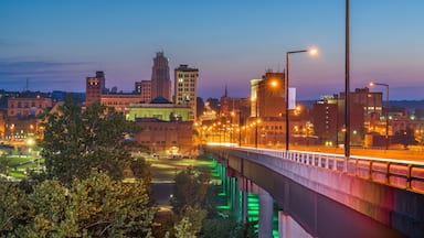 Paisagem urbana ao anoitecer, com edifícios iluminados, uma ponte e um rio.
