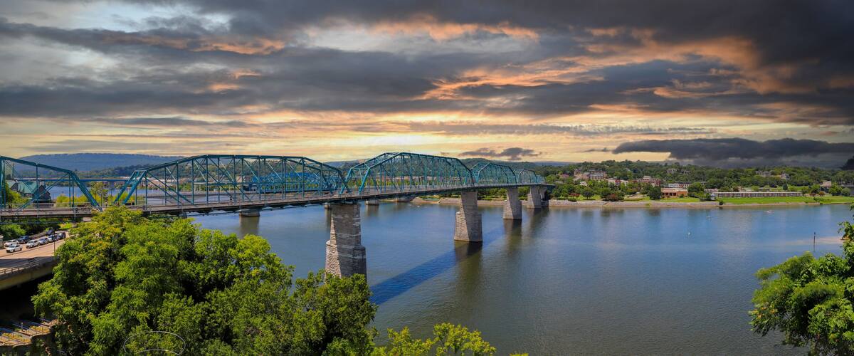 a panoramic shot of the rippling waters of the Tennessee River with the Walnut Street Bridge over the water surrounded by lush green trees with powerful storm clouds at sunset in Chattanooga