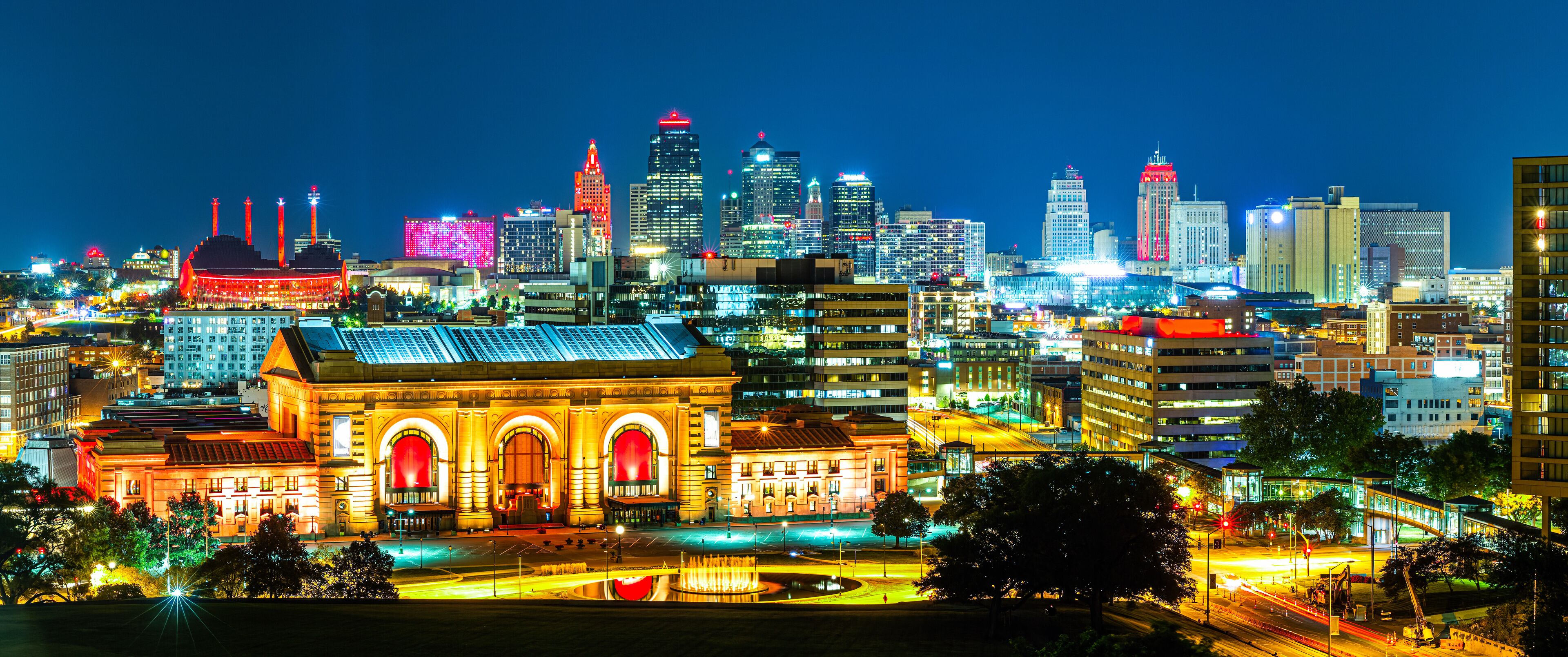 A brightly lit cityscape at night with prominent buildings, including a large, illuminated building with arched windows and a dome structure.