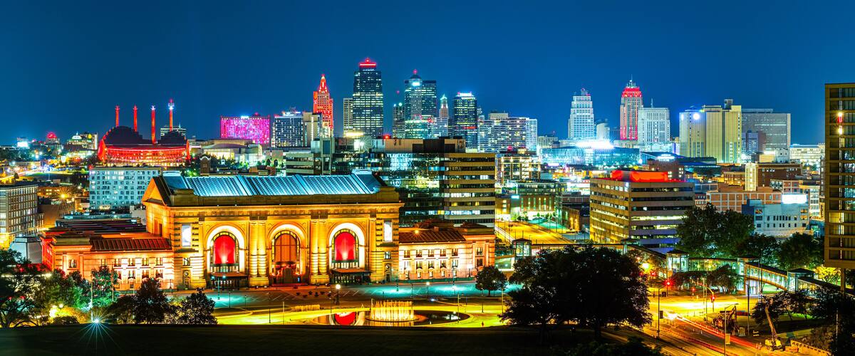 A brightly lit cityscape at night with prominent buildings, including a large, illuminated building with arched windows and a dome structure.