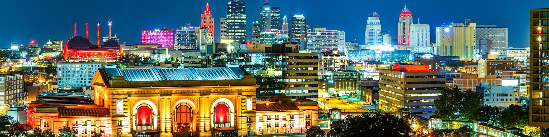 A brightly lit cityscape at night with prominent buildings, including a large, illuminated building with arched windows and a dome structure.