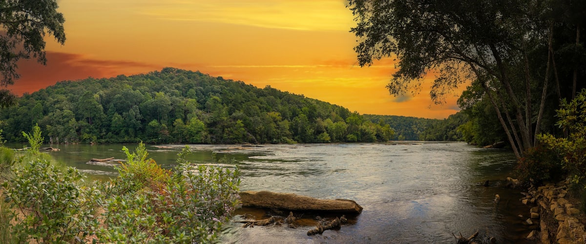 a panoramic of a gorgeous summer landscape on the Chattahoochee River with flowing water surrounded by lush green trees, grass and plants and powerful clouds at sunset in Atlanta Georgia USA